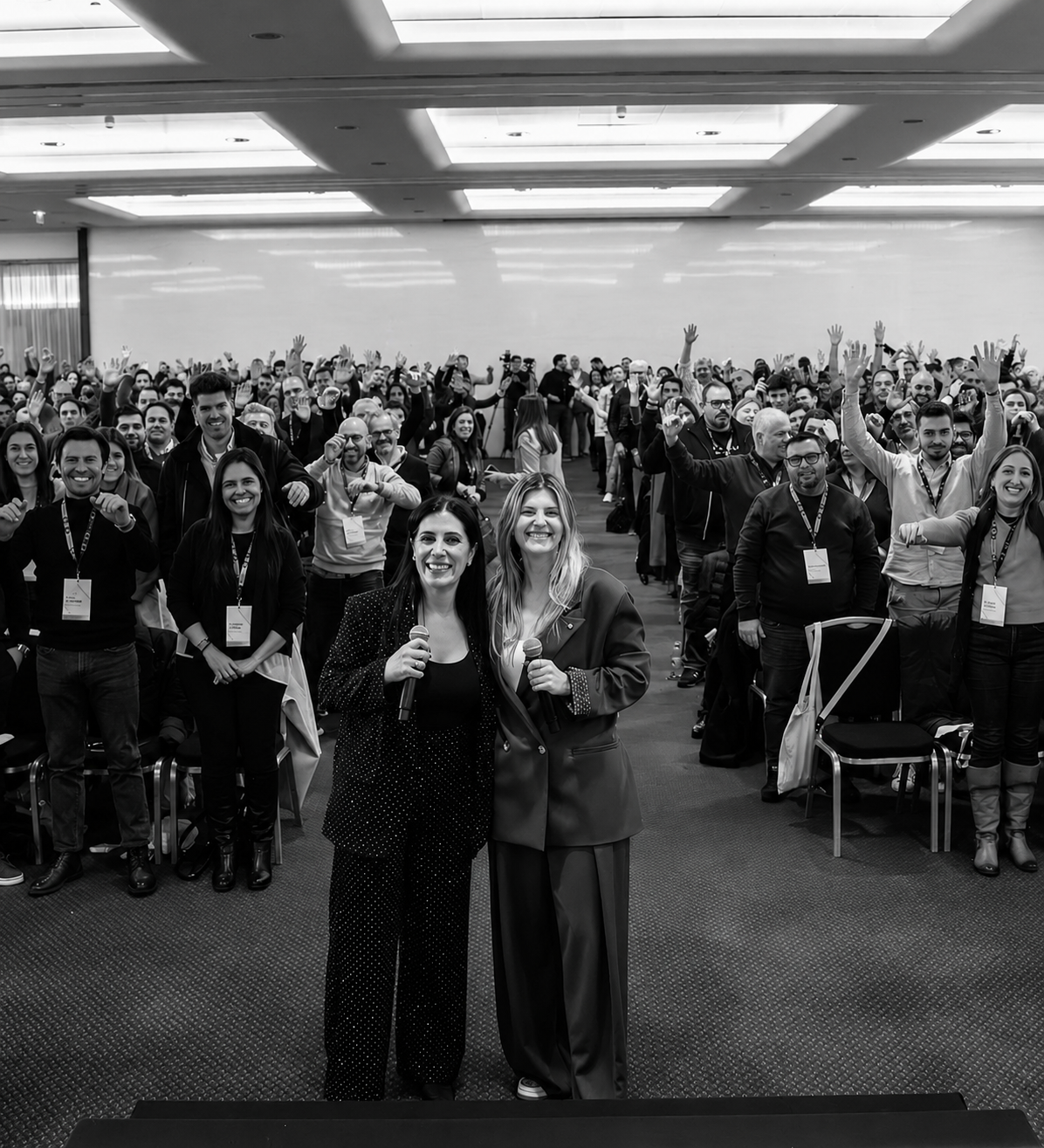Cátia and Júlia on stage with a packed audience raising hands behind them.