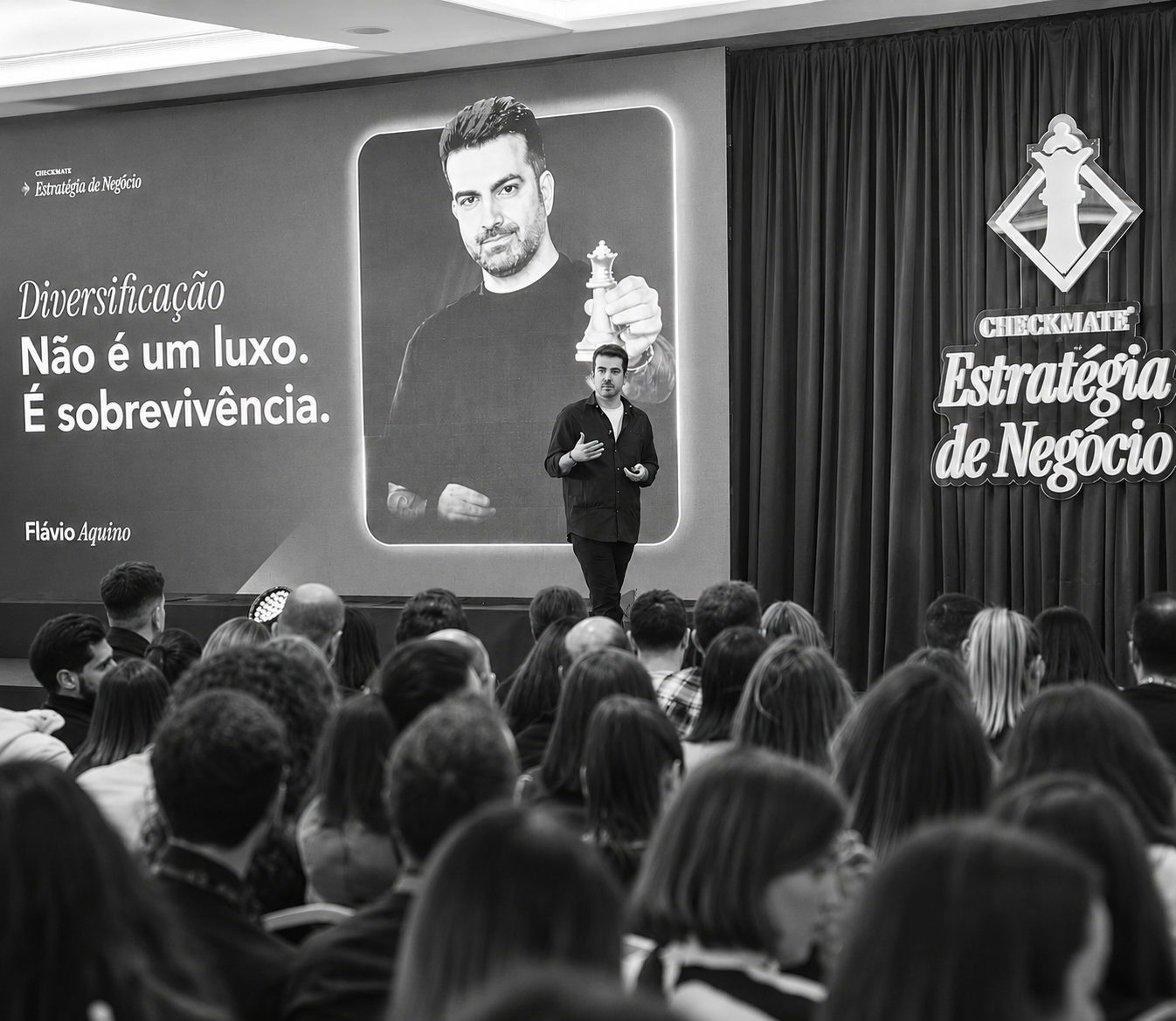 Flávio on stage at Checkmate event with a large slide reading 'Diversificação não é um luxo. É sobrevivência.'