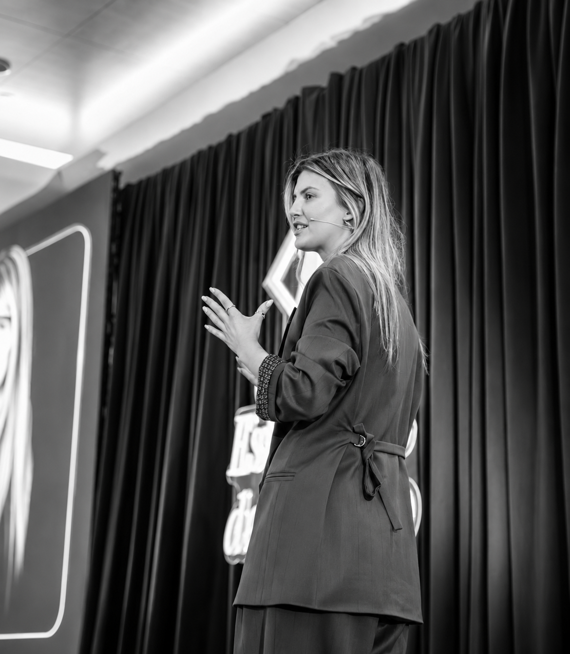 Cátia speaking on stage, side profile with stage curtain behind.